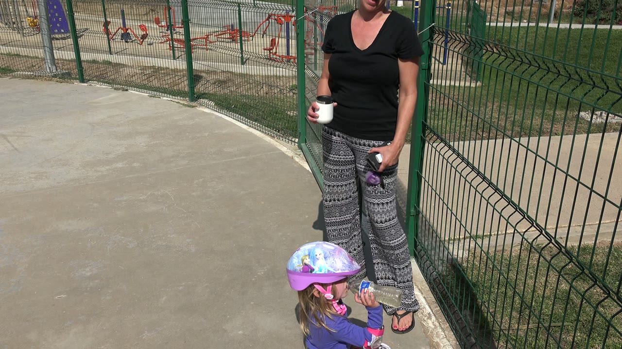 In Los Cabos, Mexico, a woman in a black shirt holds a white mug while a helmeted child drinks from a bottle near a green fence and paved walkway.
