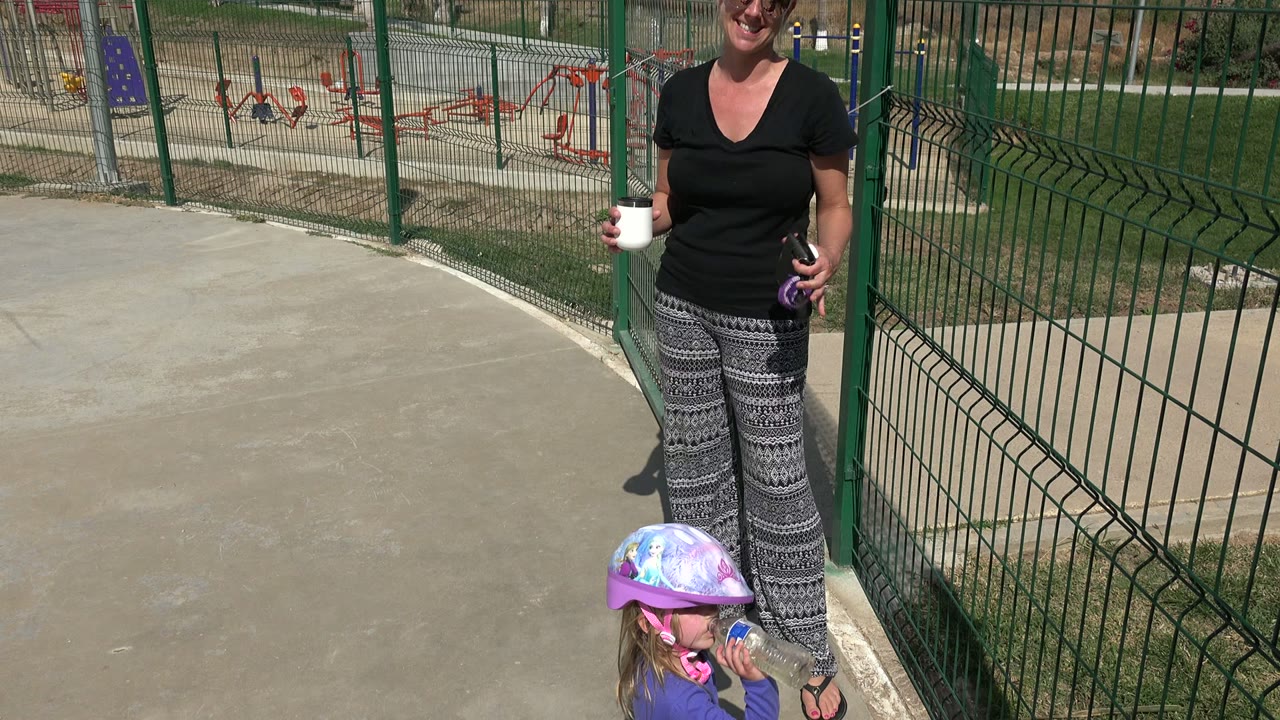 In Los Cabos, Mexico, the woman stands near the green fence with a mug in one hand as the child remains low in frame drinking from a water bottle.