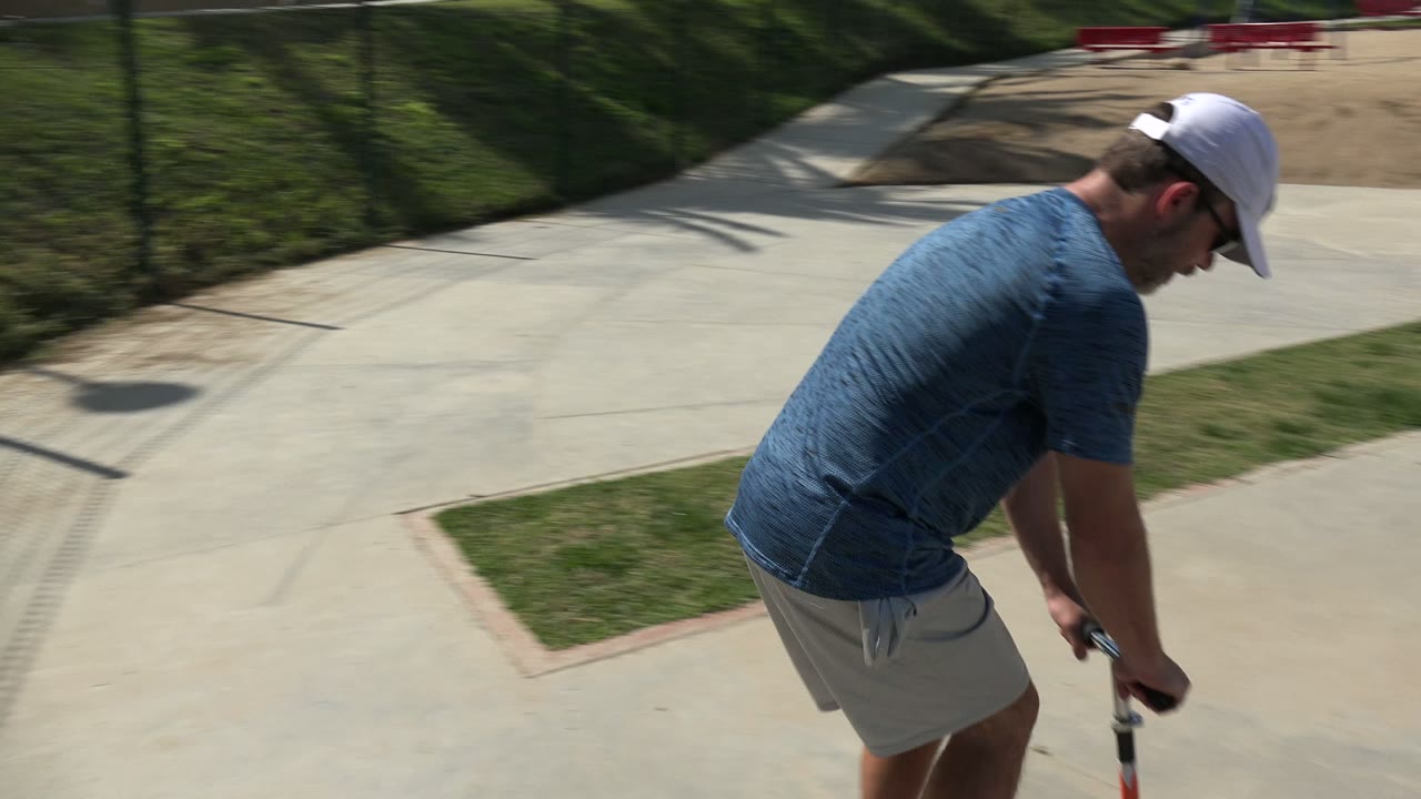 In Los Cabos, the scooter rider moves farther along the concrete path with grass strips, fence shadows, and playground sand visible ahead.