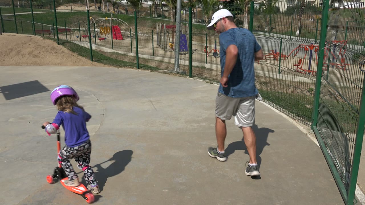 In Los Cabos, Mexico, a helmeted child rides an orange scooter behind an adult man on a concrete path beside fenced playground equipment.