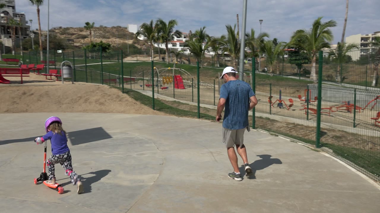 The child continues riding the scooter across a wider concrete area in Los Cabos, Mexico, while the adult man walks ahead near the fence.