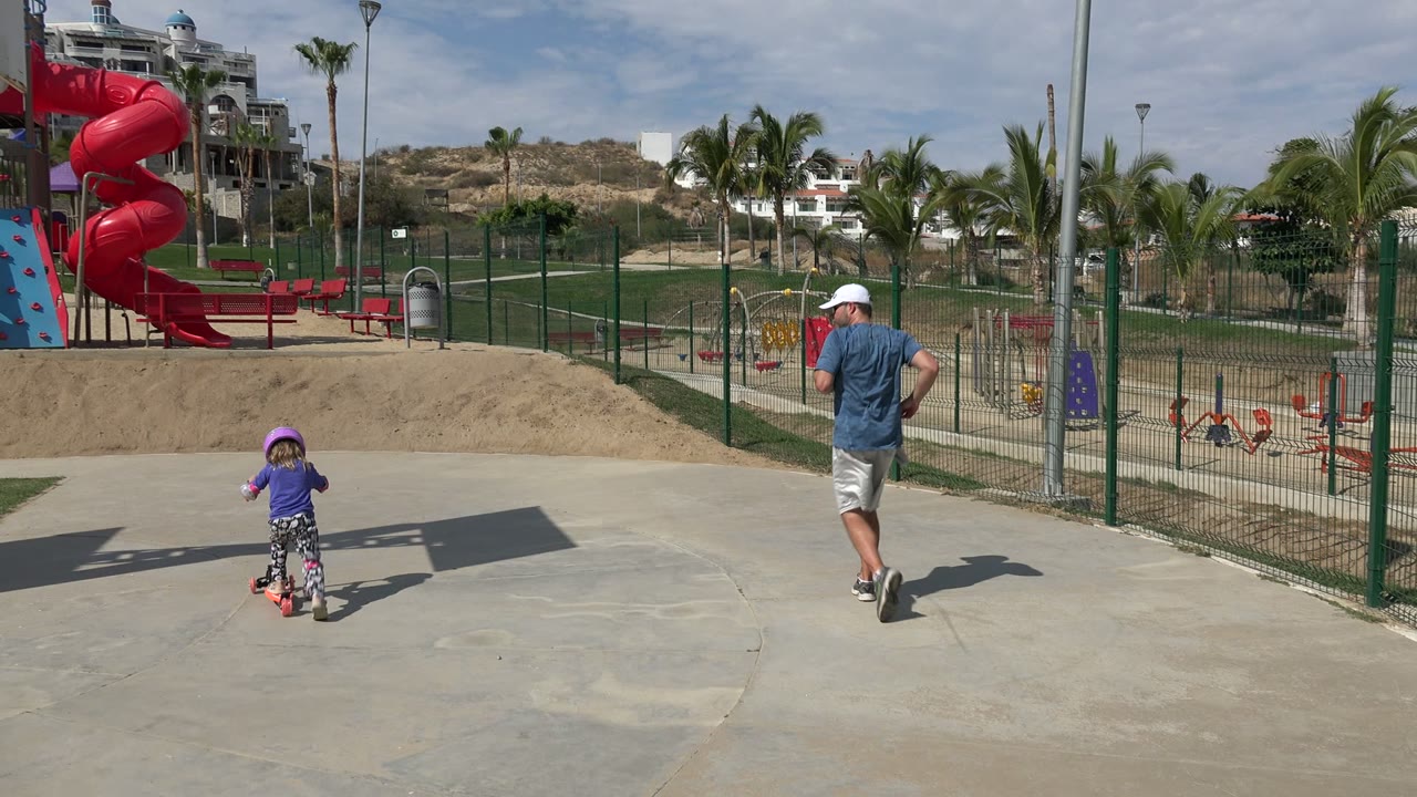 Child on scooter and adult man pass a playground with a red slide.