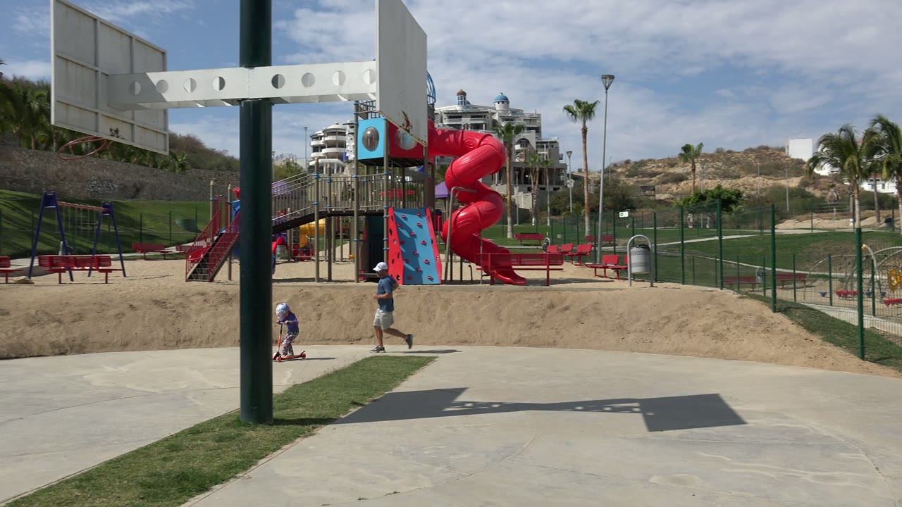One second in, the child on the scooter and the running father are farther right on the paved path, with the red spiral slide, fence, grass, palms, hillside, and distant buildings still visible in Los Cabos, Mexico.