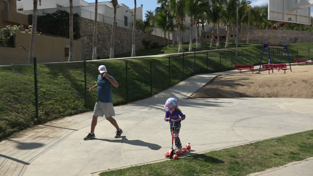 Father walks near a child riding a red scooter on a sunny park path.