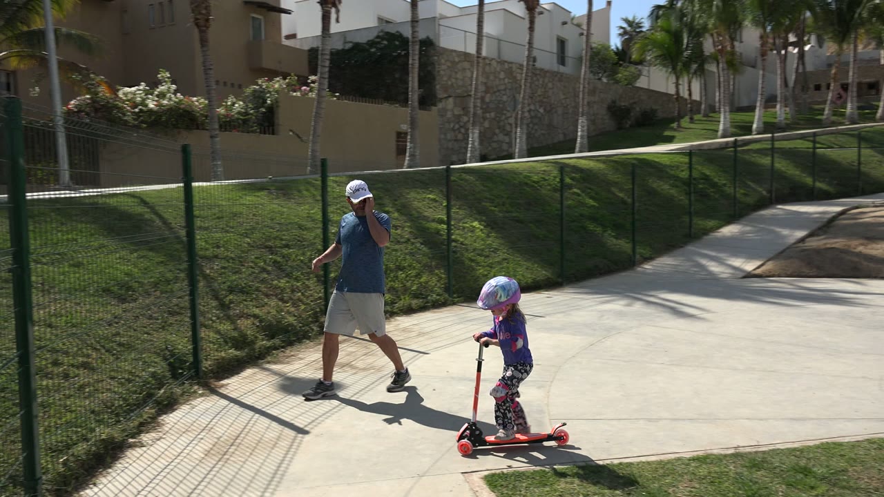 The child continues riding the red scooter along the curved walkway in Los Cabos as her father follows a few steps behind beside the fenced grass.
