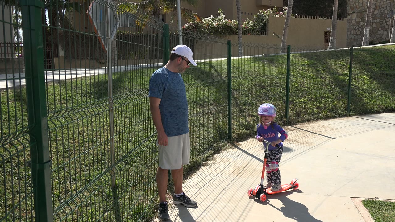Man watches a young girl standing on a red scooter beside a fence.