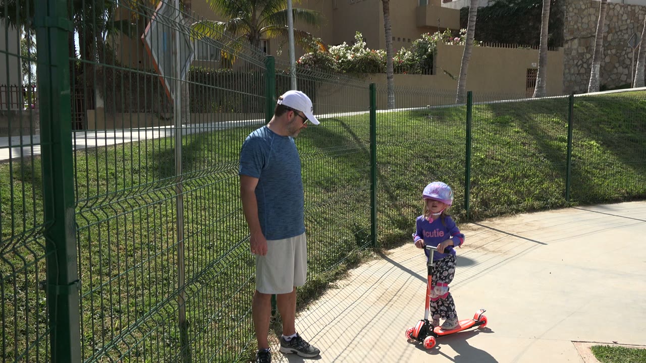 The helmeted girl rides farther along the Los Cabos sidewalk as the adult man remains near the green fence in bright sun.