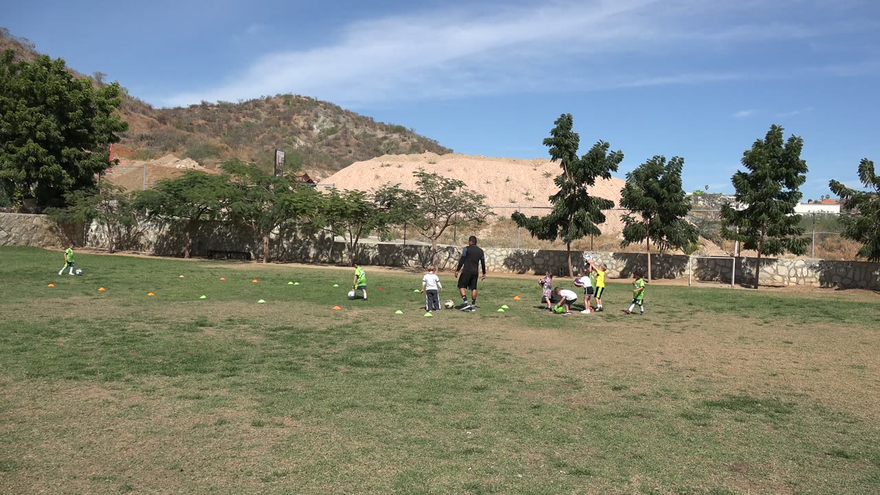 Wide view of children practicing soccer on a grass field in Los Cabos, Mexico, with an adult coach near the center, training cones across the turf, trees, a low wall, and dry hills behind them.