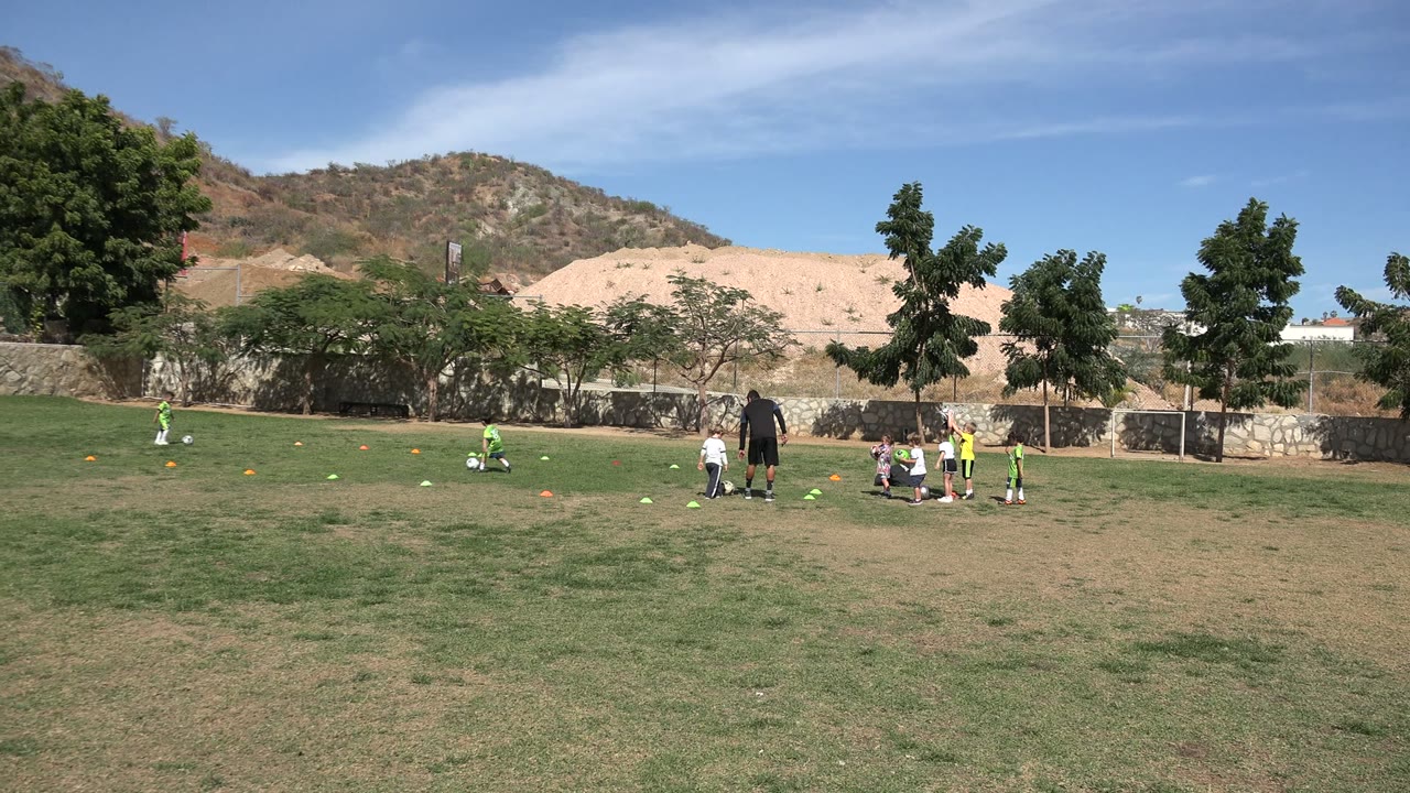 Children in green soccer uniforms continue drills on the Los Cabos grass field while the adult coach stands nearby and a seated cluster of kids gathers near several balls.