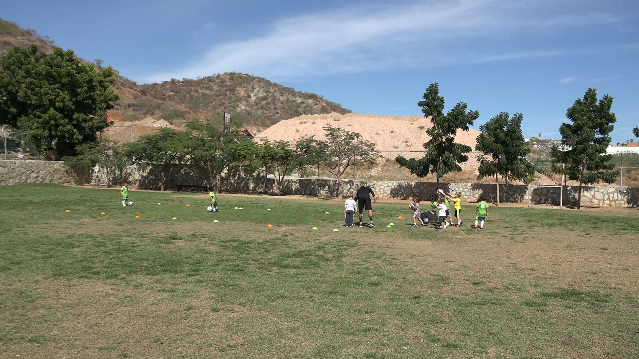 Youth soccer practice on grass with coach, cones, goal, trees, and hills.