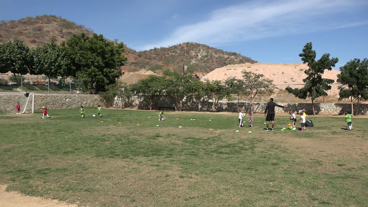 Children practice football on a grassy field with a goal and hillside behind them.