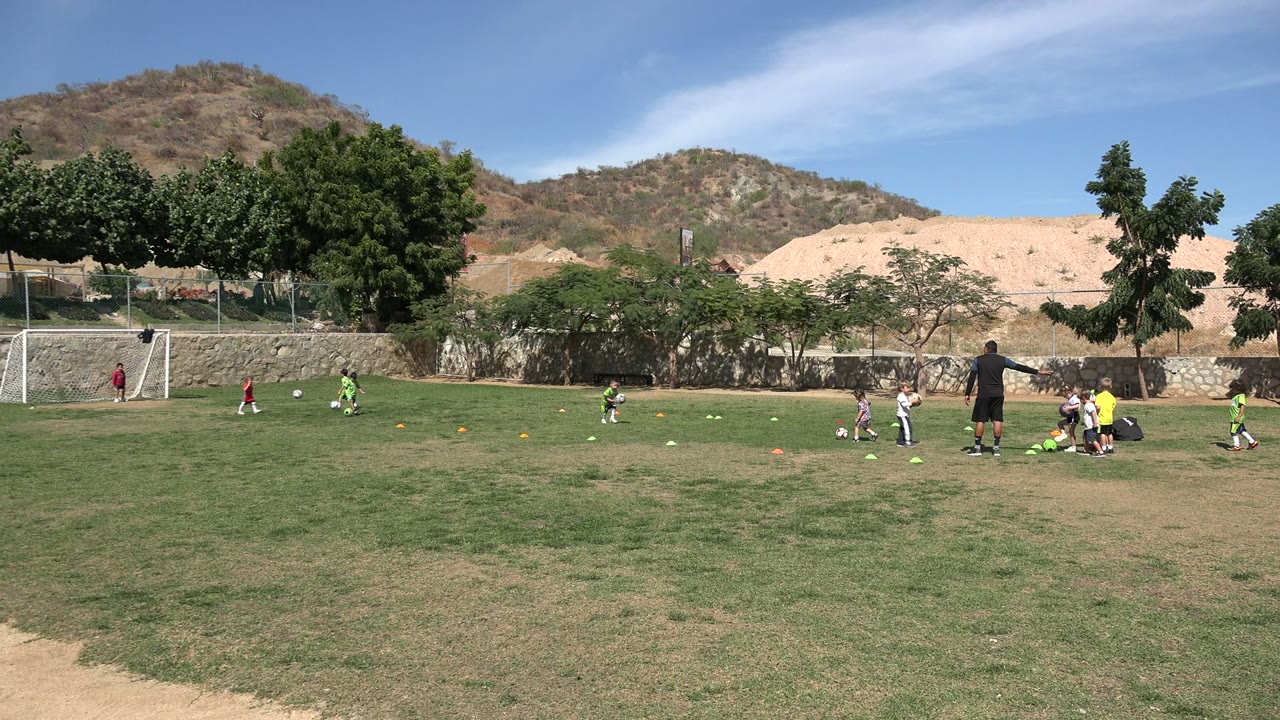Children continue a football drill in Los Cabos, Mexico, with one child near the center of the field, an adult coach watching from the right, and the goal, trees, wall, and hills behind them.