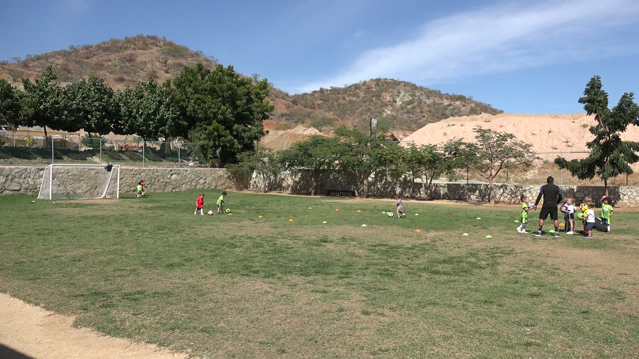 Children practice soccer on a grass field near a goal and dry hills.