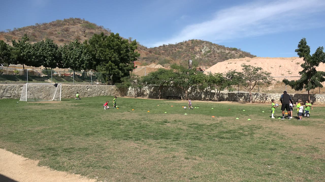 Children continue soccer drills in Los Cabos, Mexico, with one player in red near the center-left and an adult coach standing by a cluster of children on the right.