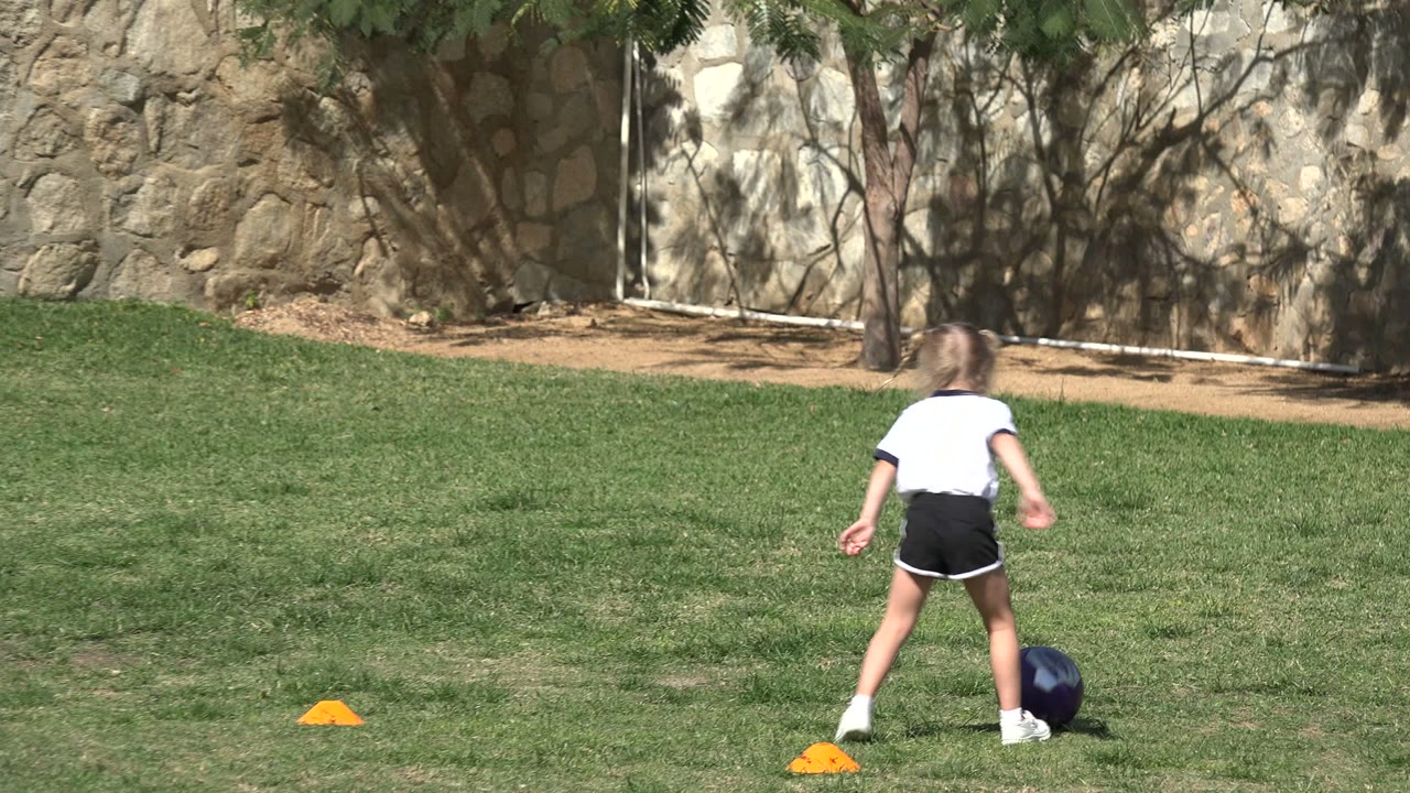 The child pauses or adjusts near the dark soccer ball in Los Cabos, Mexico, with orange cones spaced across the grass and tree shadows on the stone wall in back.