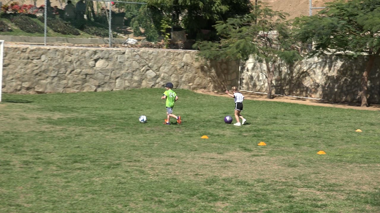 A child in a bright green shirt dribbles a football across the grass in Los Cabos, Mexico, while another child stands near a purple ball and cones mark the practice area.
