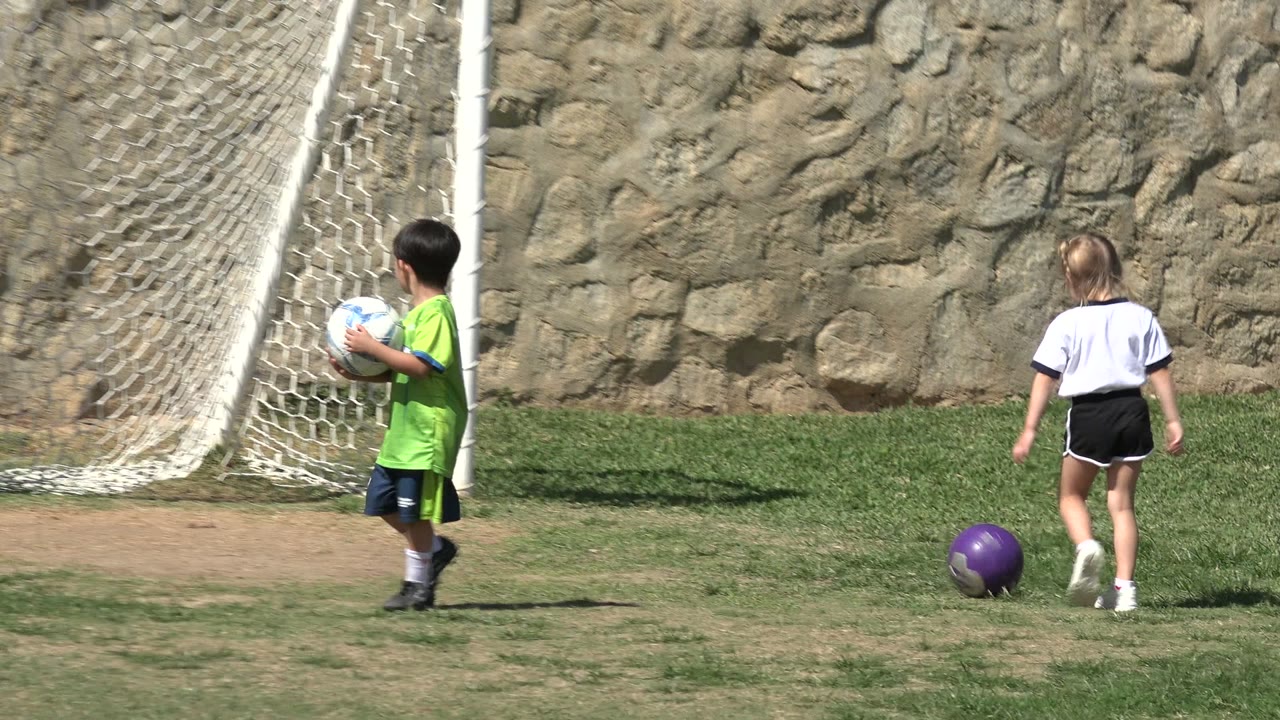 In Los Cabos, Mexico, a boy in a green soccer jersey holds a white soccer ball near a net while a girl in a white jersey walks across the grass.
