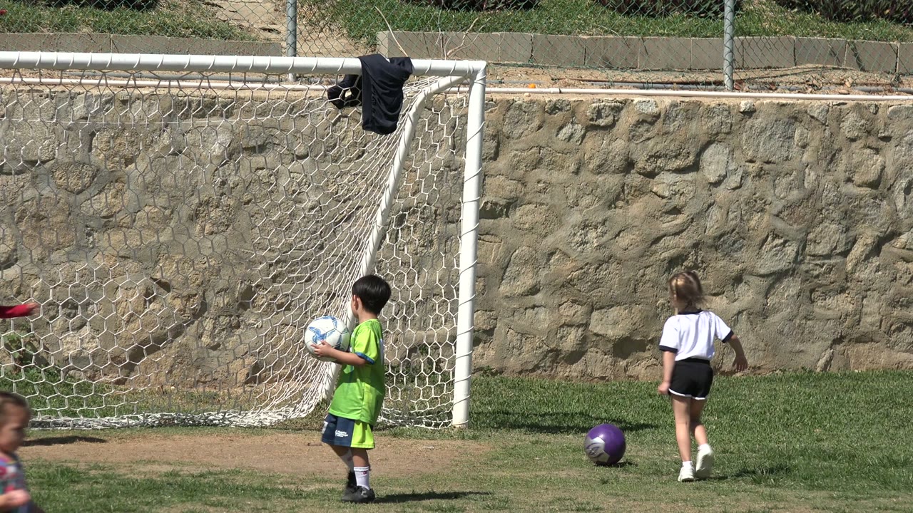 In Los Cabos, Mexico, the child in a green jersey stands in front of the soccer goal holding the ball as a girl in white moves farther right.