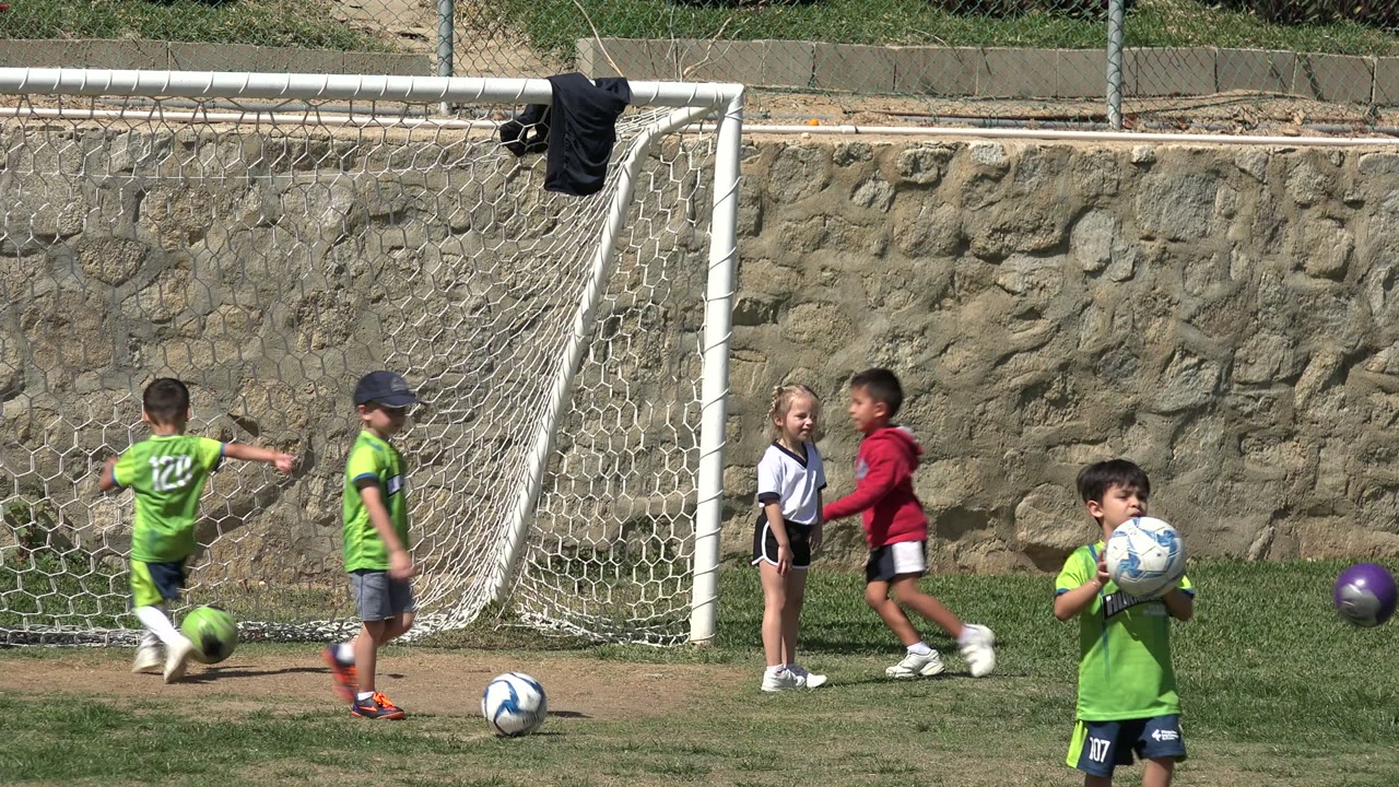 Young children in green jerseys and casual soccer clothing cluster near the goal while balls sit on the grass in Los Cabos, Mexico.