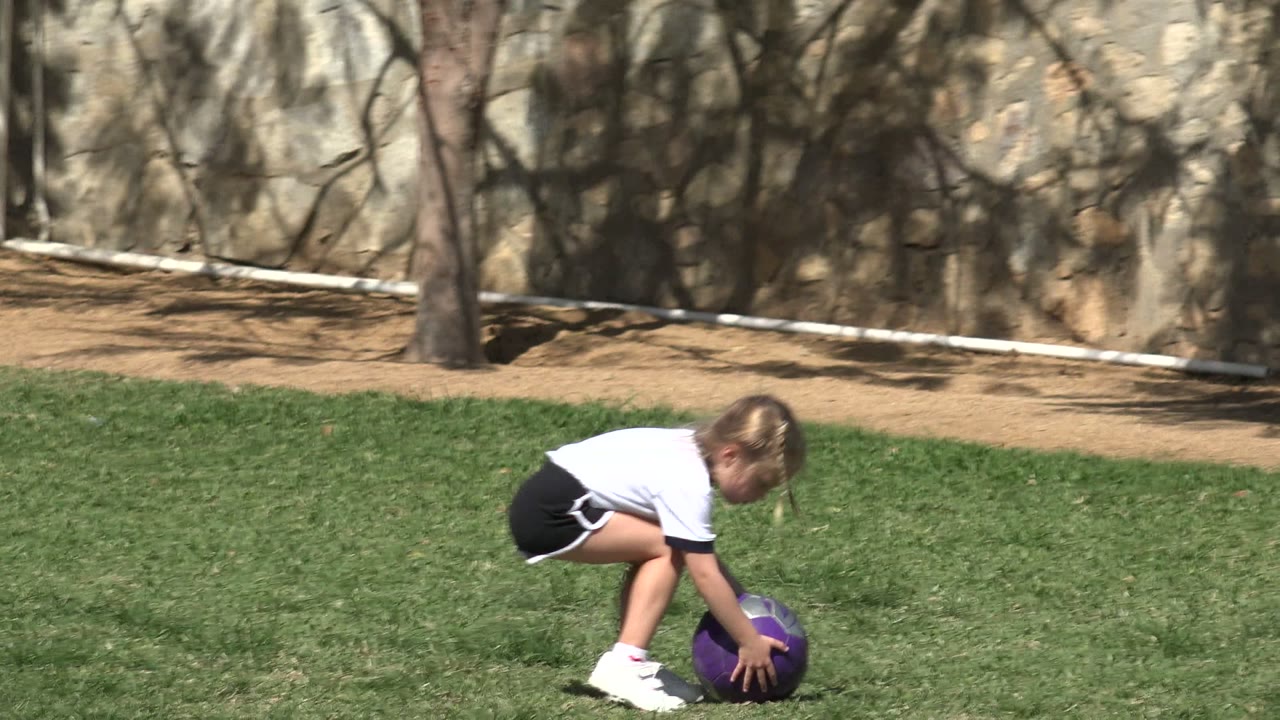 Young girl bends down to pick up a purple soccer ball on grass.