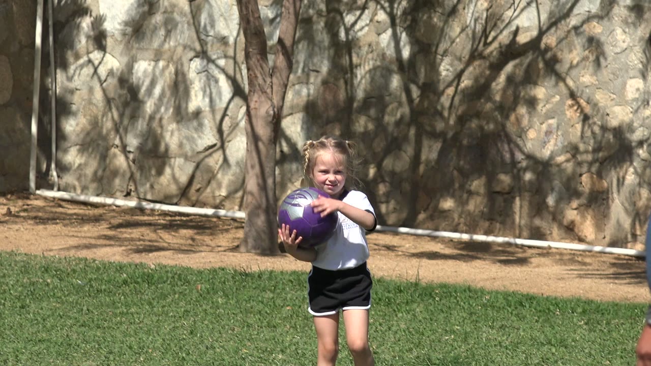 A young girl holds a purple soccer ball against her chest on the Los Cabos grass field, with tree shadows and a stone wall behind her.