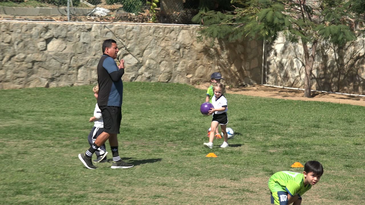 At 0:01 in Los Cabos, Mexico, the coach walks across the grass while children in green and white soccer outfits move with soccer balls nearby.