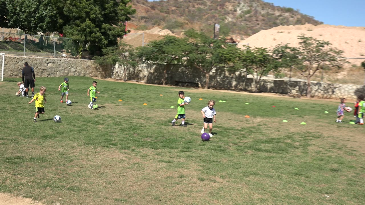 A young girl in a white jersey stands near a purple soccer ball in the foreground as other children practice soccer across a Los Cabos grass field.