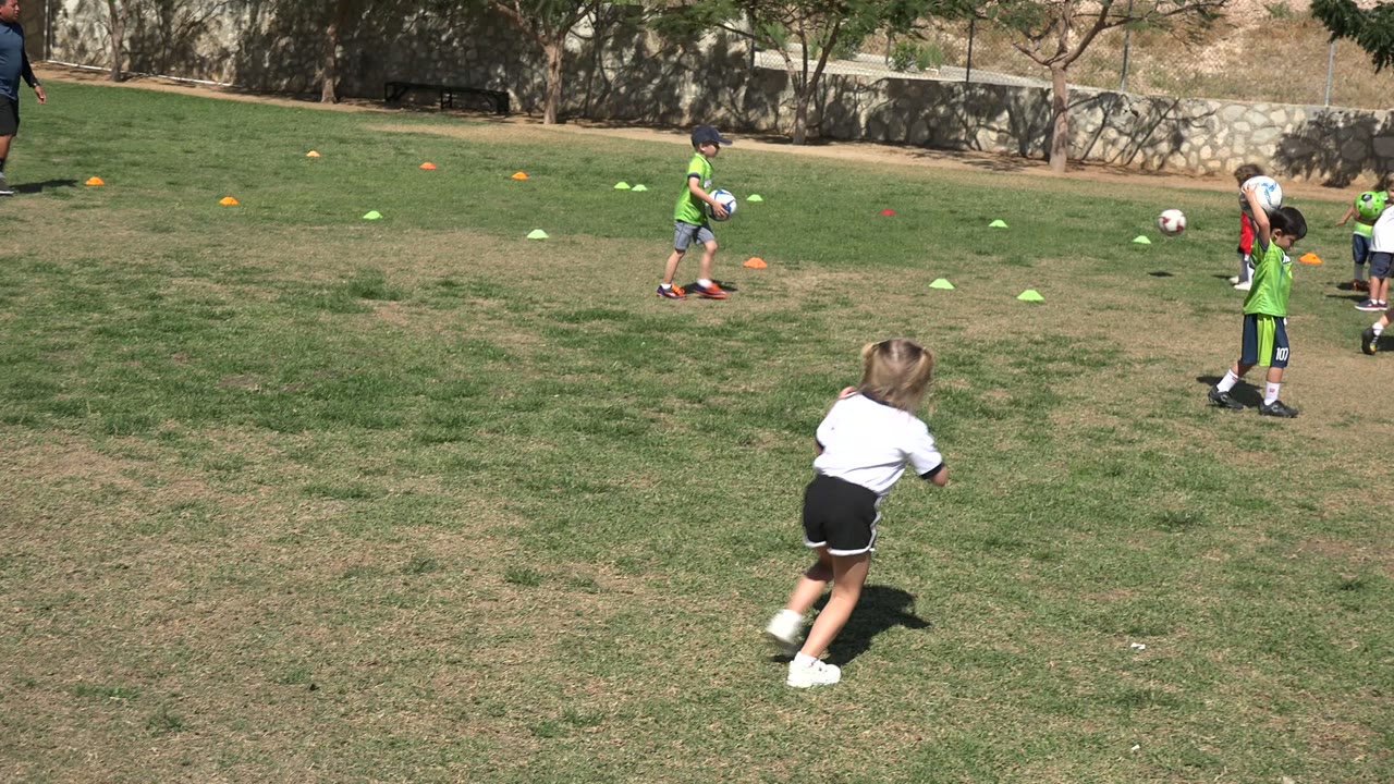 A child in a white shirt and black shorts stands near a purple ball in the foreground, while other children practice with balls farther back on the Los Cabos field.