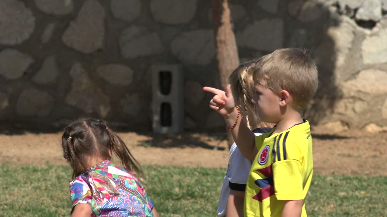 A boy in a yellow soccer jersey points across the grass in Los Cabos while two other children stand close beside him near the stone wall.
