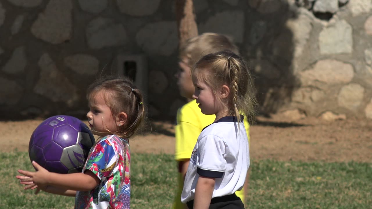 Two girls stand near a purple ball as a boy in yellow passes behind them.