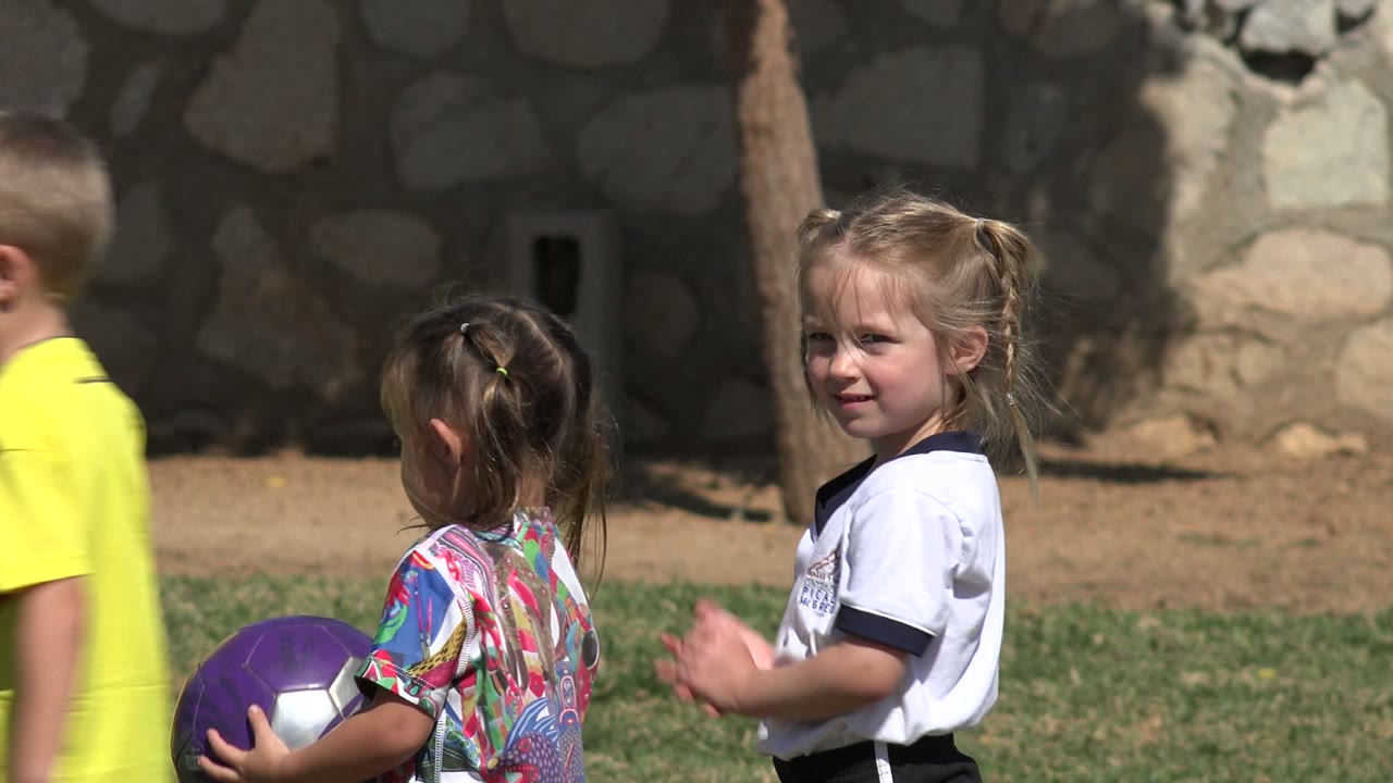In Los Cabos, Mexico, a young girl in a colorful shirt holds the purple ball as another girl in a white shirt faces her on the sunny field.
