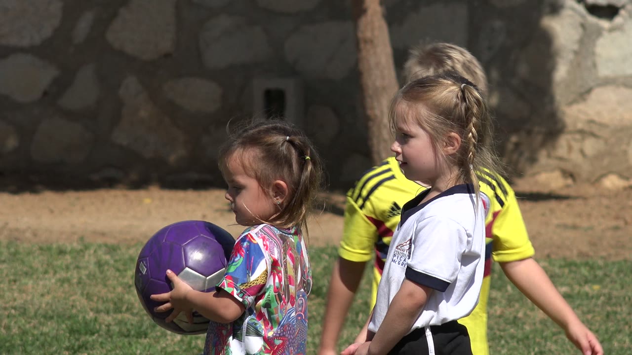 In Los Cabos, Mexico, a small girl holds a purple soccer ball while two other children stand nearby on a sunlit grass field.