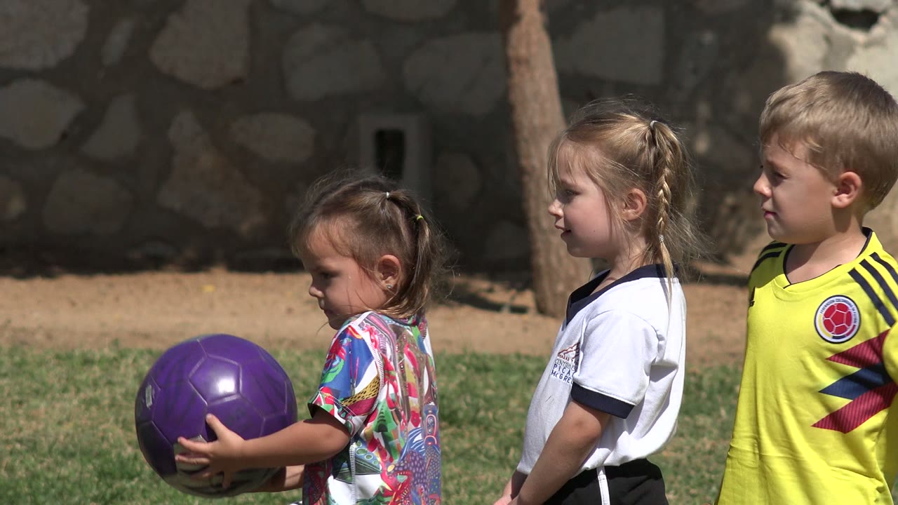 Three children in soccer clothing stand in profile on a grassy field in Los Cabos, Mexico, with the purple ball held low at the left edge.
