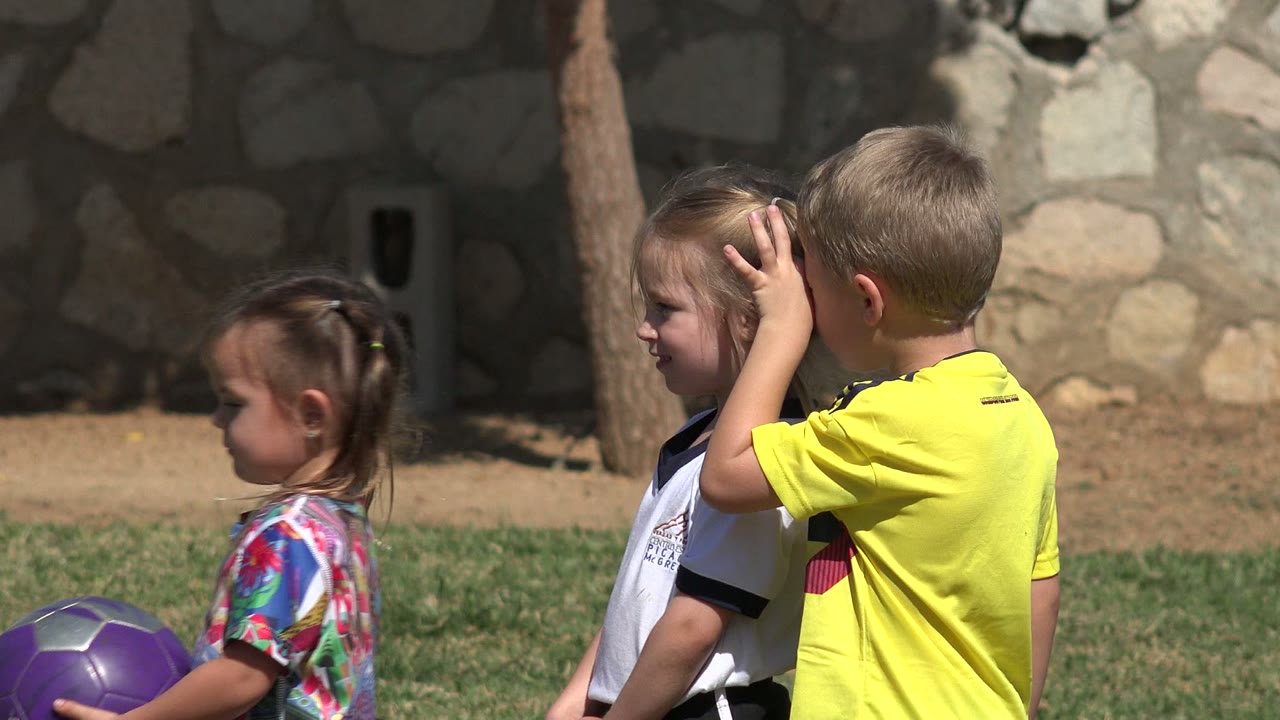 One second into the Los Cabos, Mexico footage, a boy in a bright yellow shirt holds his hand up beside a girl in a white soccer jersey while another child with a purple ball stands nearby.