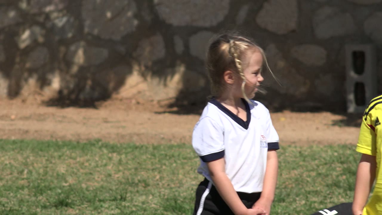 The girl faces to the side on the Los Cabos soccer field while part of another child in a yellow jersey appears at the edge of the frame.