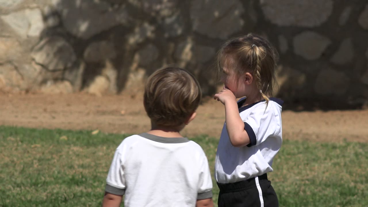 Two children in white shirts stand on grass near a stone wall.