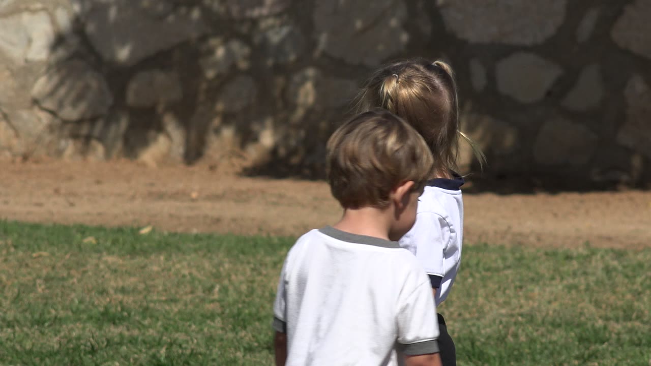 Two children in white shirts face across the grass in Los Cabos, Mexico, while a third child is partly obscured behind them against a sunlit stone wall.