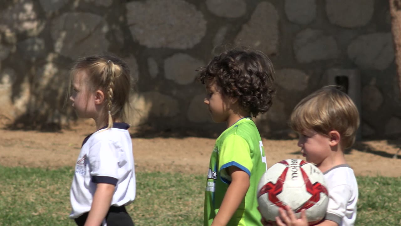 Three young children on a grass field near a stone wall.