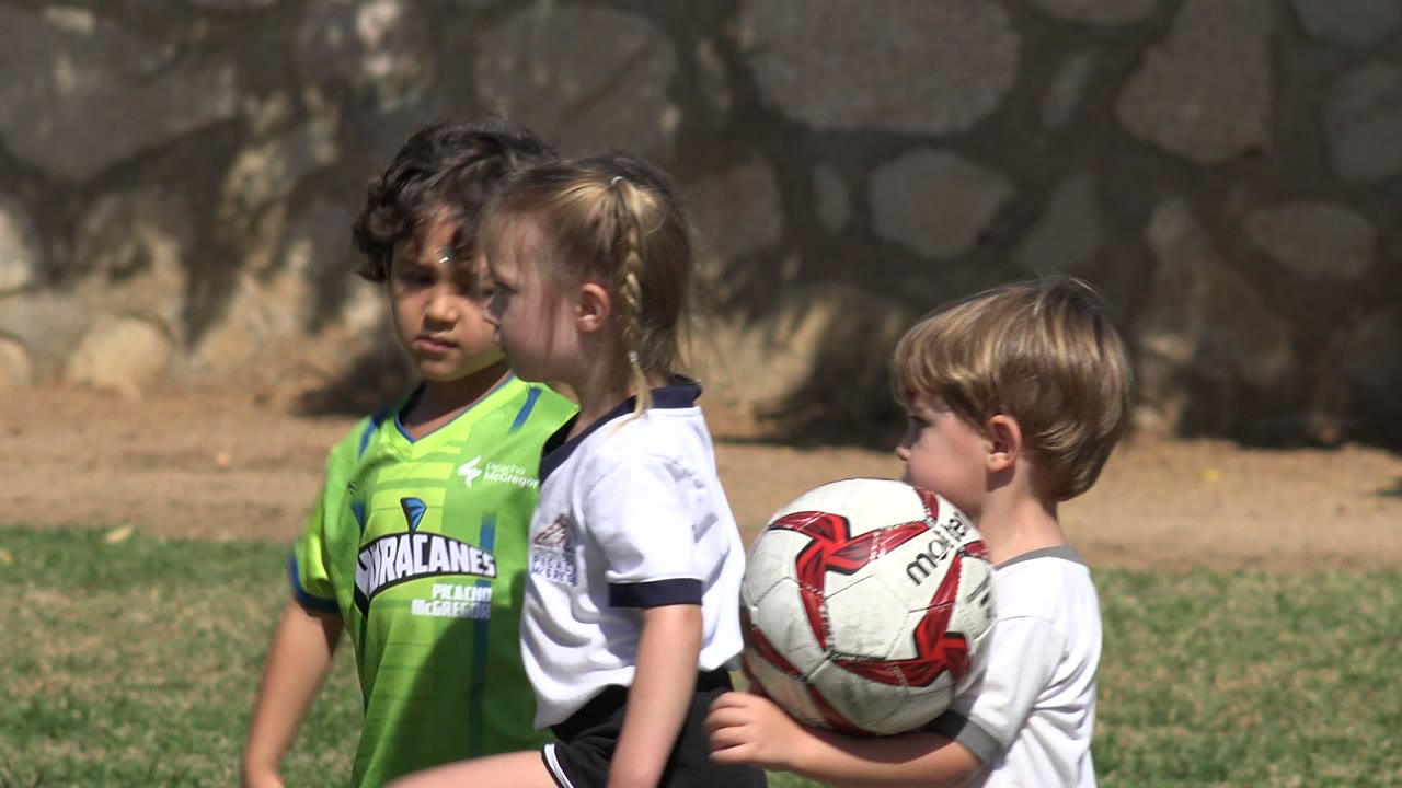 A child in a green soccer shirt faces the camera while two children in white shirts stand close together on the Los Cabos grass field, with part of the soccer ball entering the foreground.