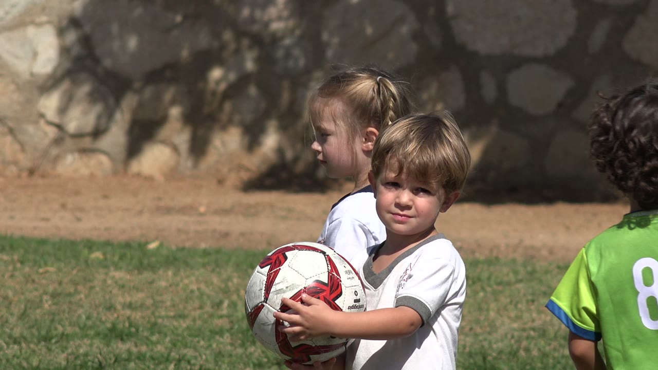 At the start of the Los Cabos, Mexico clip, a young boy in a white shirt holds a soccer ball while other children in green and white soccer jerseys stand nearby on the grass.