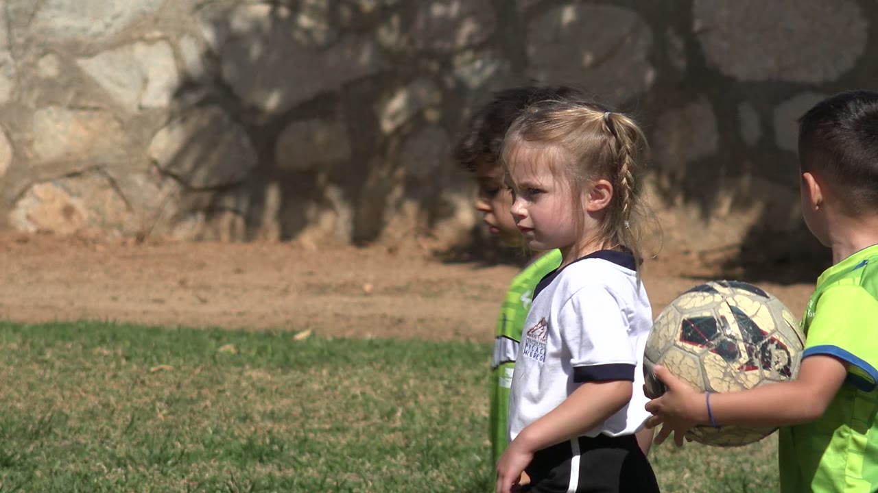 Young children in soccer shirts holding balls on grass near a stone wall.