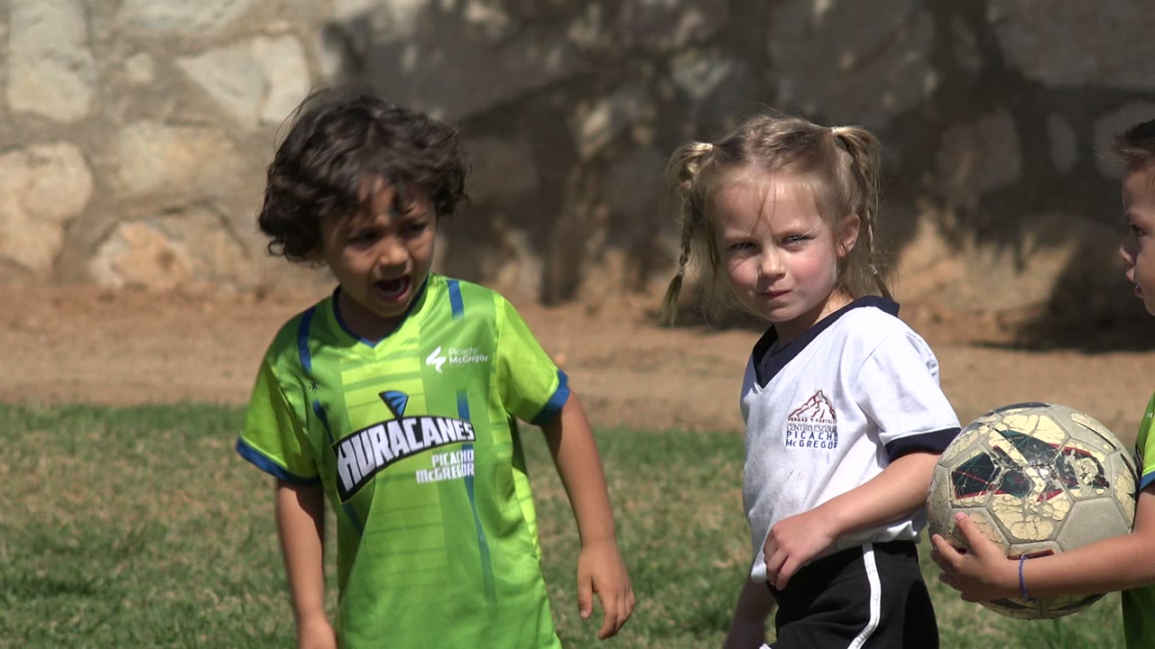 A child in a lime green soccer jersey looks toward the camera area in Los Cabos, Mexico, while another child nearby holds a scuffed soccer ball on the grass.