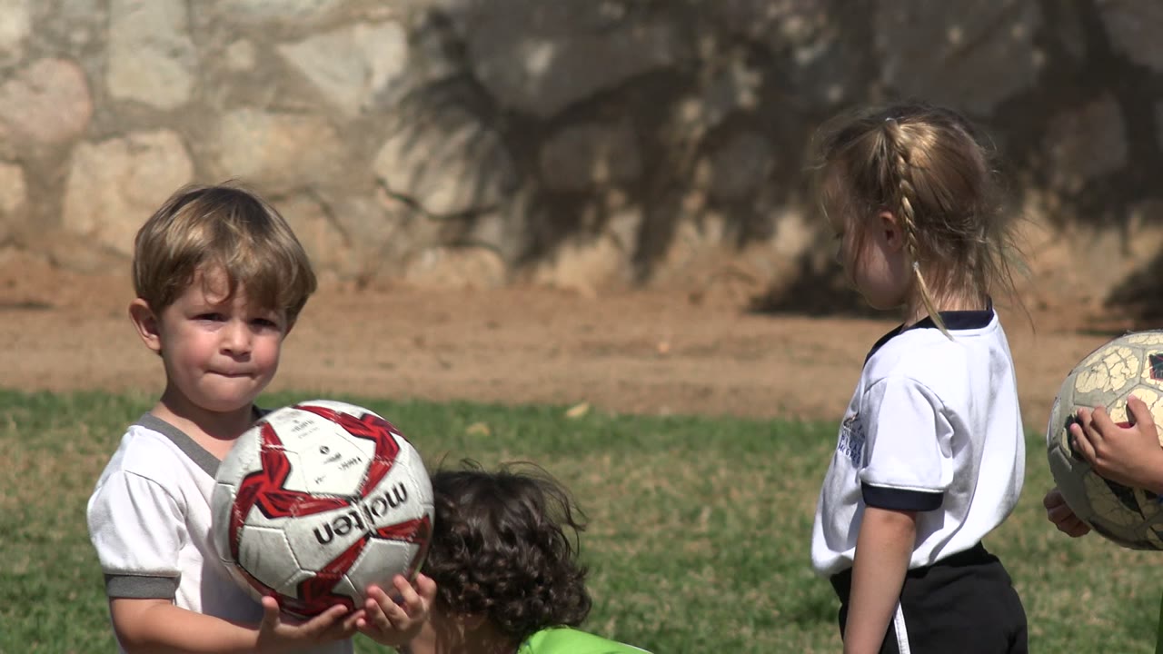 Young boy holding a soccer ball beside other children on a grass field.