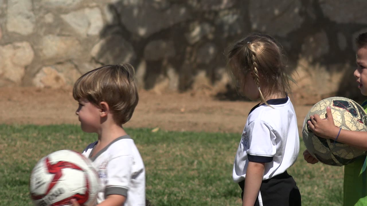 In Los Cabos, Mexico, a blonde girl in a white shirt stands in profile as a young boy holds a worn soccer ball in front of her on the field.