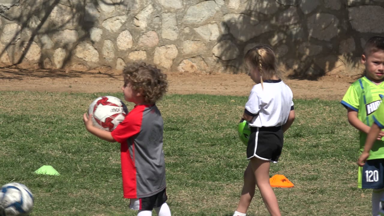 A young child in a red and gray jersey holds a soccer ball on the Los Cabos field while a girl in white and a green-jersey player stand nearby in bright sun.