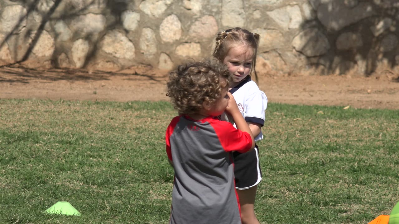 Two children stand on a sunny grass field in Los Cabos, Mexico, with a red-and-gray shirt in the foreground, a white shirt facing the camera, green cones on the grass, and a stone wall behind them.