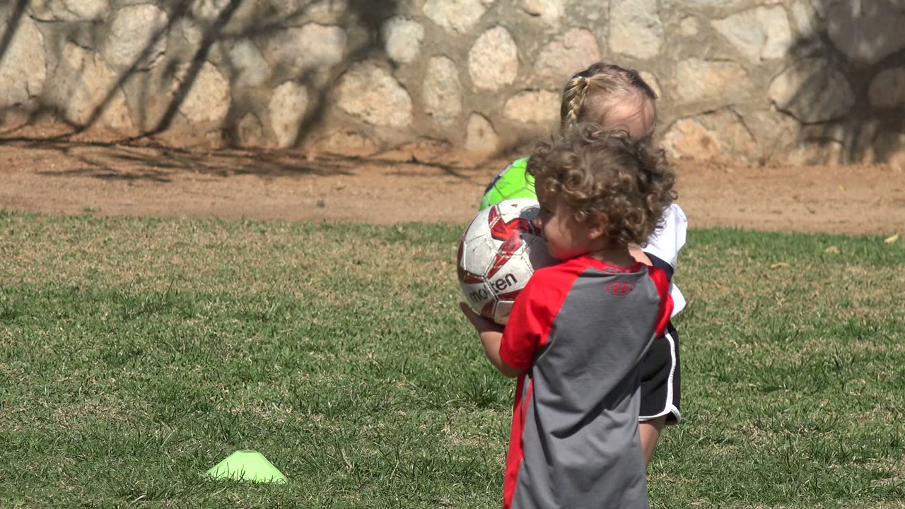 Children on a Los Cabos grass field gather around two soccer balls, with a curly-haired child seen from behind and a girl in a white shirt partly hidden near the stone wall background.