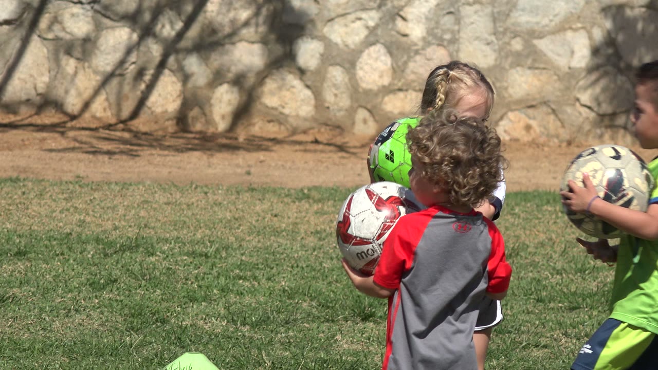 Children hold soccer balls while standing in bright sun on grass.