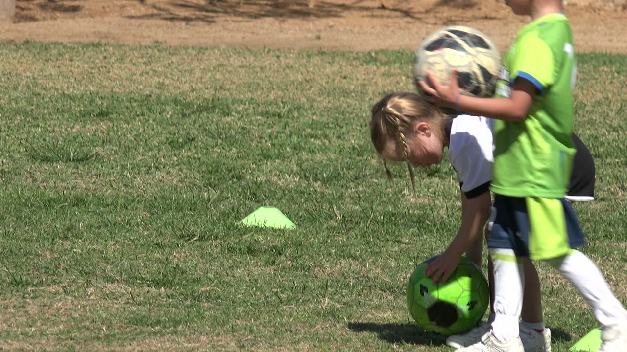 A child in a green soccer kit holds a ball while a girl in a white shirt bends toward a bright green ball on the grass in Los Cabos, Mexico.