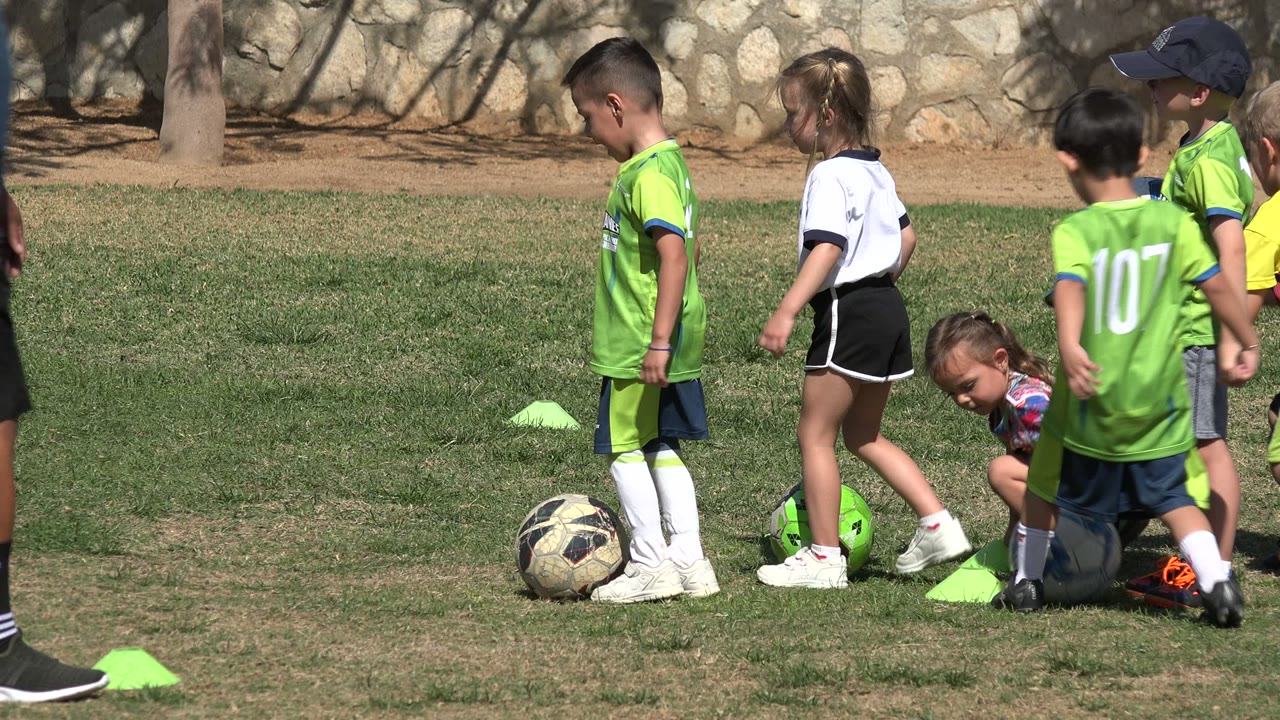 Children in soccer uniforms move around balls and bright cones on the Los Cabos grass field, with one child crouched near a ball.